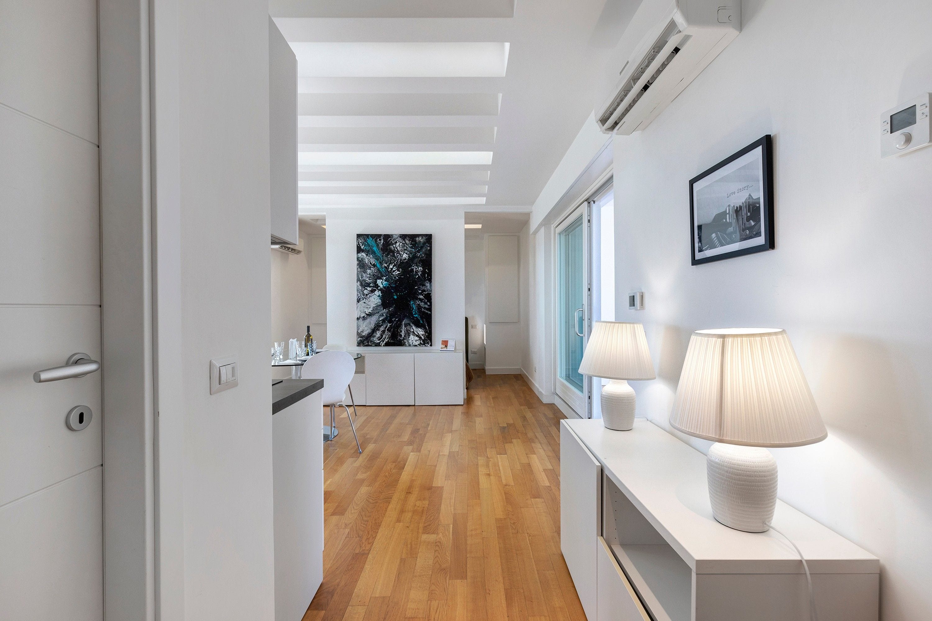 Un couloir moderne avec un plancher en bois, une console blanche agrémentée de lampes et un tableau encadré accroché au mur.