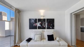Un salon moderne avec un canapé blanc, trois tableaux abstraits et une porte-fenêtre coulissante donnant sur un balcon.