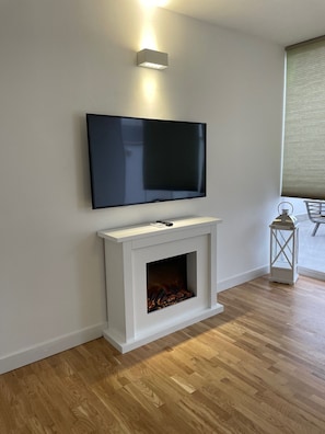 Un soggiorno moderno con una TV a schermo piatto montata sopra un caminetto bianco, un pavimento in legno e una finestra con persiane.