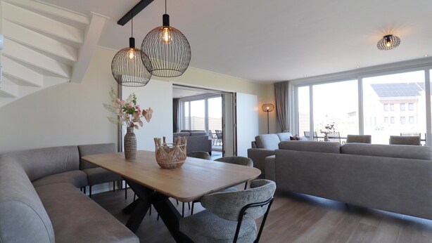 A modern living room with a sofa, a dining table, and hanging pendant lights. - Newly Built Group Villa in Friesland (Leons)