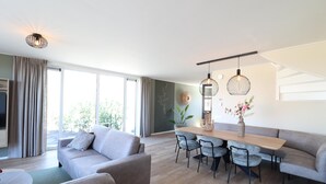 A modern living room with a sofa, dining table, and chairs, a large window with curtains, and a hanging light fixture. - Newly Built Group Villa in Friesland (Leons)