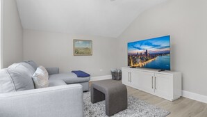 A modern living room with a grey sofa, a small ottoman, a white cabinet, and a large TV displaying a cityscape. - Elegant 1 Bed Steps to Metra & Dining in Lake Forest! (Lake Forest)