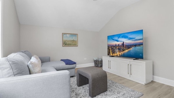 Una sala de estar moderna con un sofá gris, un pequeño reposapiés, un mueble blanco y un televisor grande que muestra un paisaje urbano.