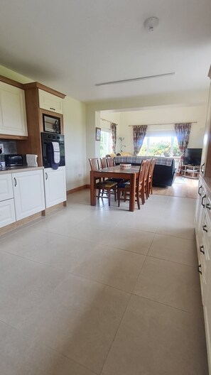 A spacious living area with a dining table and chairs, a kitchen with white cabinets, and a large window providing natural light. -  An ideal base for families and couples looking to explore the area. (Duncormick)