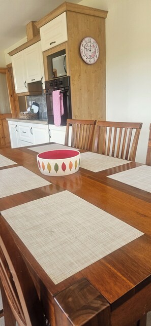 A dining table with placemats and chairs, a clock on the wall, and a kitchen area with cabinets and an oven. -  An ideal base for families and couples looking to explore the area. (Duncormick)
