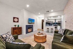 Ein Wohnzimmer mit Kamin, grünen Sofas, einem runden Couchtisch und einer Küche im Hintergrund.