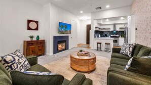 Ein Wohnzimmer mit Kamin, grünen Sofas, einem runden Couchtisch und einer Küche im Hintergrund.