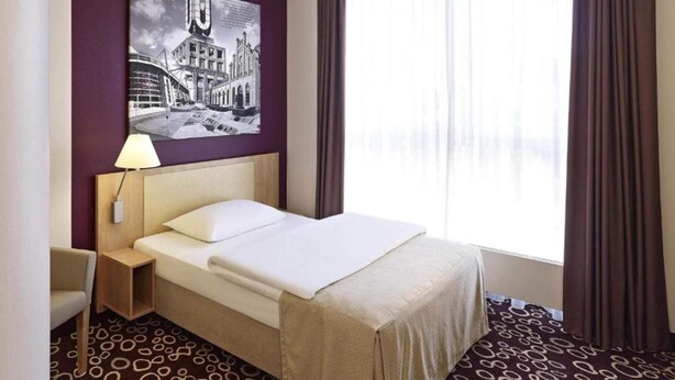 A hotel room with a bed, a nightstand, a lamp, a large window with curtains, and a wall-mounted picture. - 1 Person Holiday Park Home in Dortmund (Dortmund)