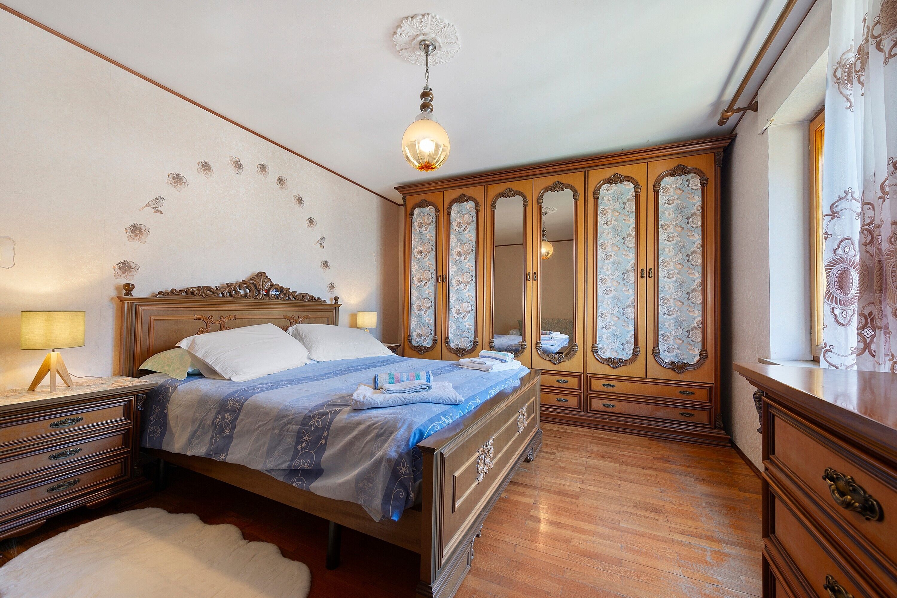 Ein Schlafzimmer mit einem hölzernen Bett, einem Nachttisch, einer Kommode und einem großen Spiegel mit floralem Dekor.