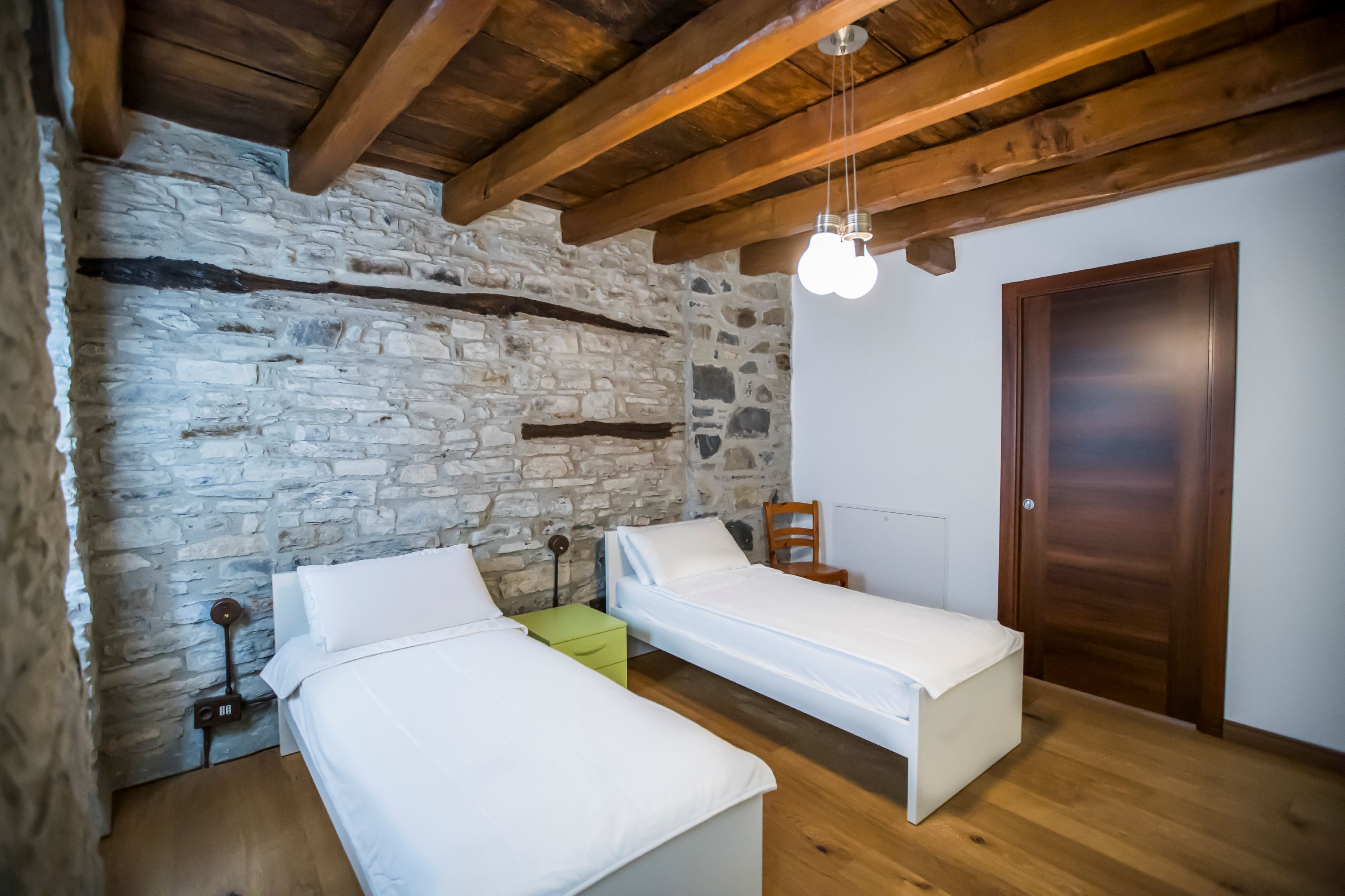 Quarto com duas camas de solteiro, parede de pedra, vigas de madeira, porta de madeira e luminária pendente.