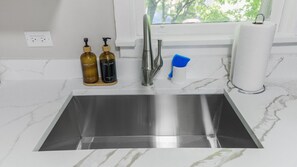 Een keuken met een spoelbak van roestvrij staal, twee zeepdispensers, een kraan, een houders voor keukenpapier en een raam met uitzicht op groen.