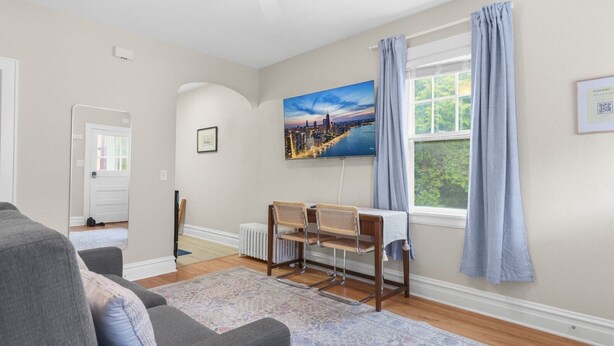 Un salon comprenant un canapé, une table avec des chaises, un radiateur, une fenêtre avec des rideaux et un tableau représentant un paysage urbain accroché au mur.