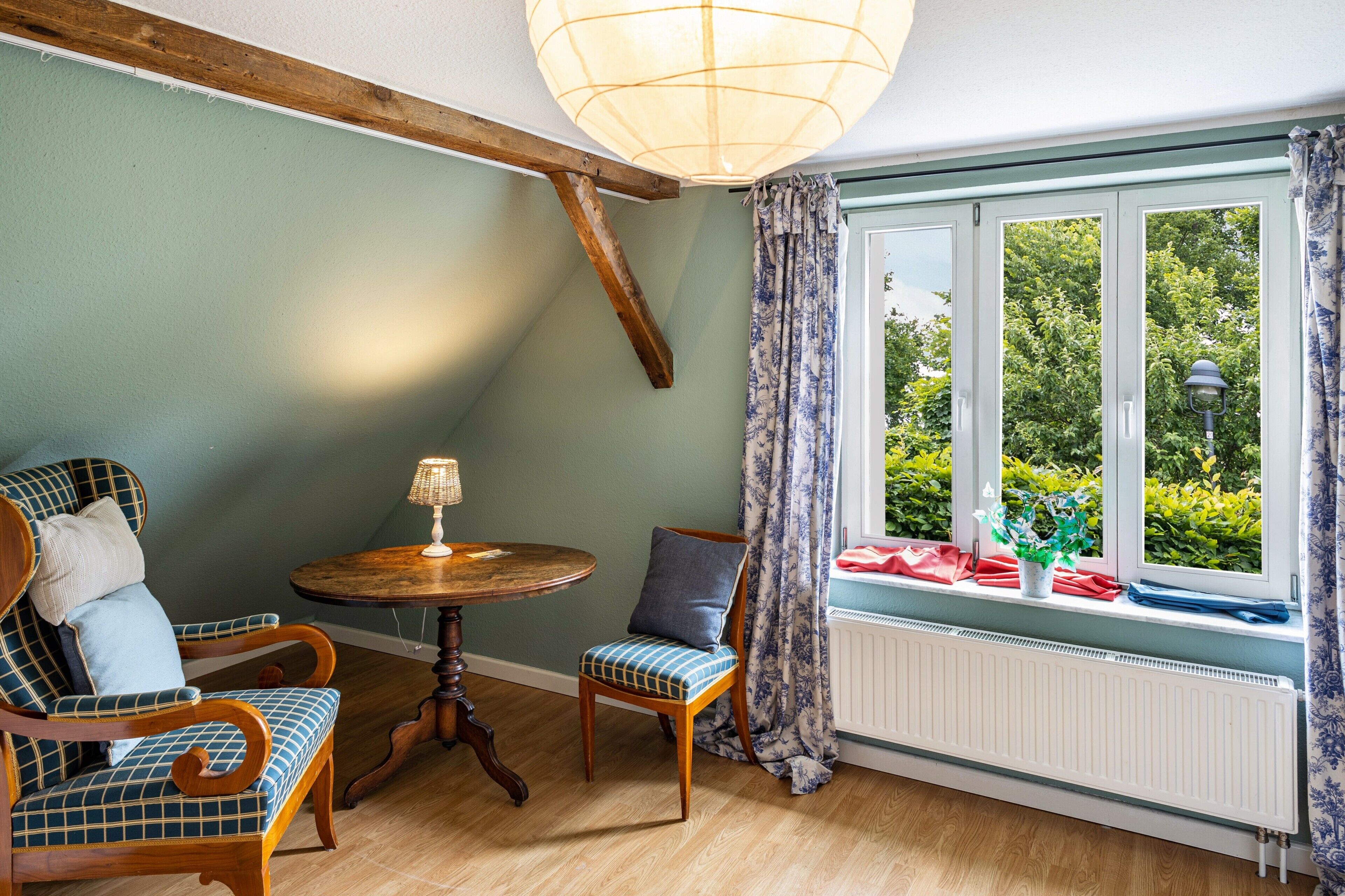A room with a wooden ceiling beam, a window with a view of greenery, a table with a lamp, and two chairs with patterned upholstery.
