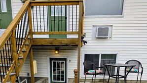 Ein zweistöckiges Haus mit einer Holztreppe, einer grünen Tür und einer Terrasse mit zwei Stühlen und einem Tisch.