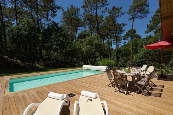A wooden deck with a pool, lounge chairs, and a table with chairs. - Grande villa au cœur de la forêt avec piscine (Moliets)