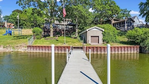 A dock leading to a small building with a garage door, surrounded by greenery and a playground. - Charming 3 Bed Lakefront Home steps to the water! (Wonder Lake)