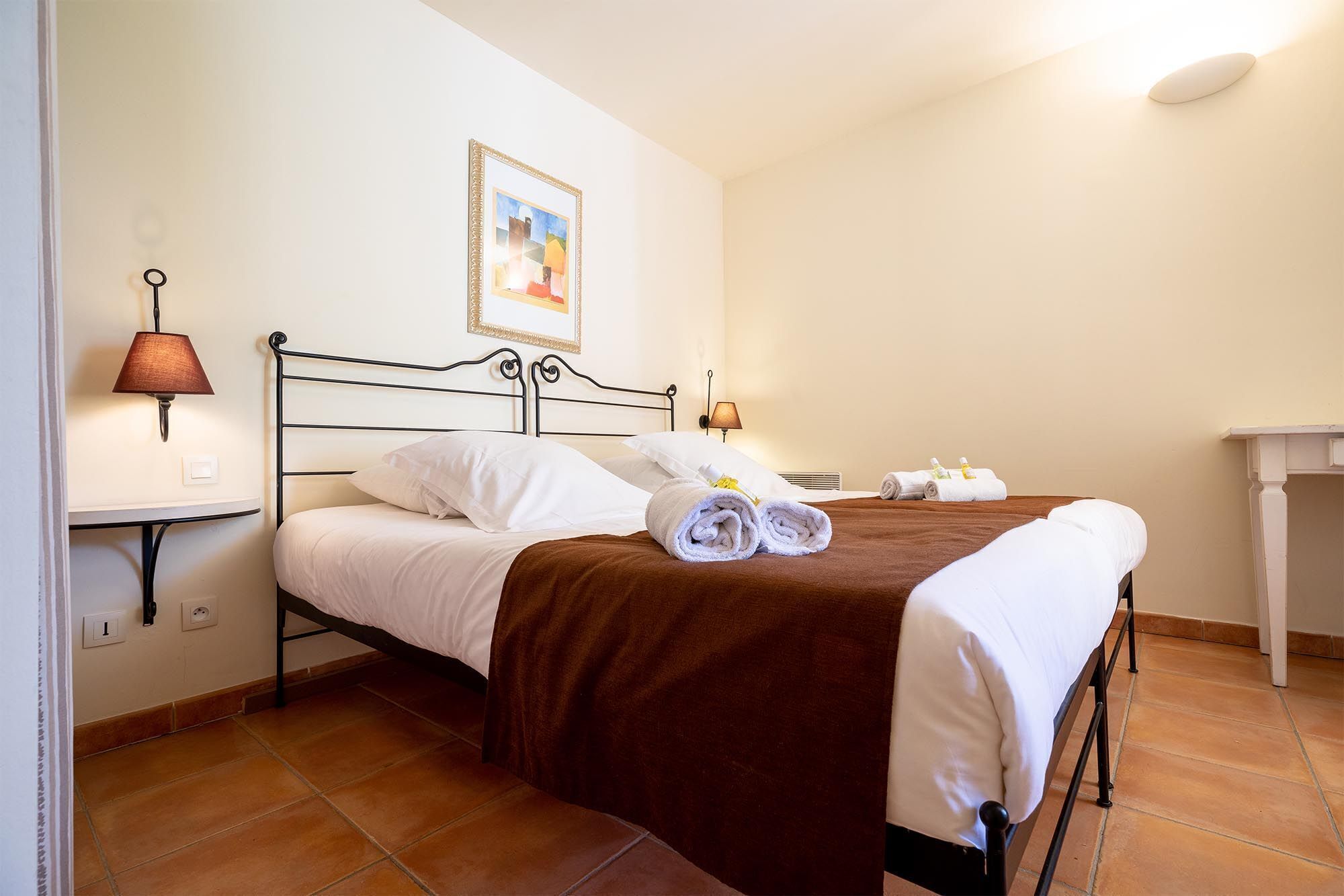 A neatly made bed with white linens, a brown throw blanket, and rolled towels on a metal frame bed in a room with tiled flooring and a small table with toiletries.