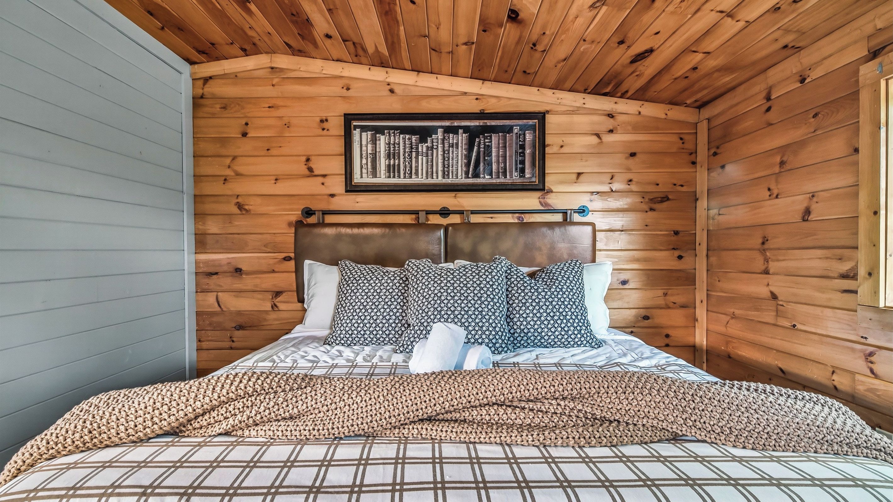 A wooden-paneled room with a bed, a framed book collection on the wall, and a window.