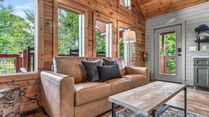A wooden cabin interior with a leather sofa, a wooden table, and a view of the outdoors.