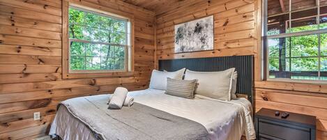 A wooden cabin bedroom with a bed, two windows, a headboard, and a nightstand.