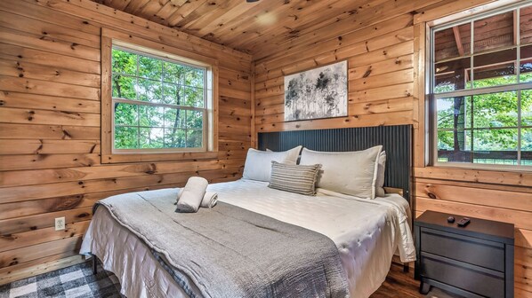 A wooden cabin bedroom with a bed, two windows, a headboard, and a nightstand.