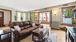 A living room with a brown leather sofa, a wooden coffee table, and a view of the outdoors through large windows. - Walk to Downtown Sturgeon Bay! Large Home + Yard (Sturgeon Bay)