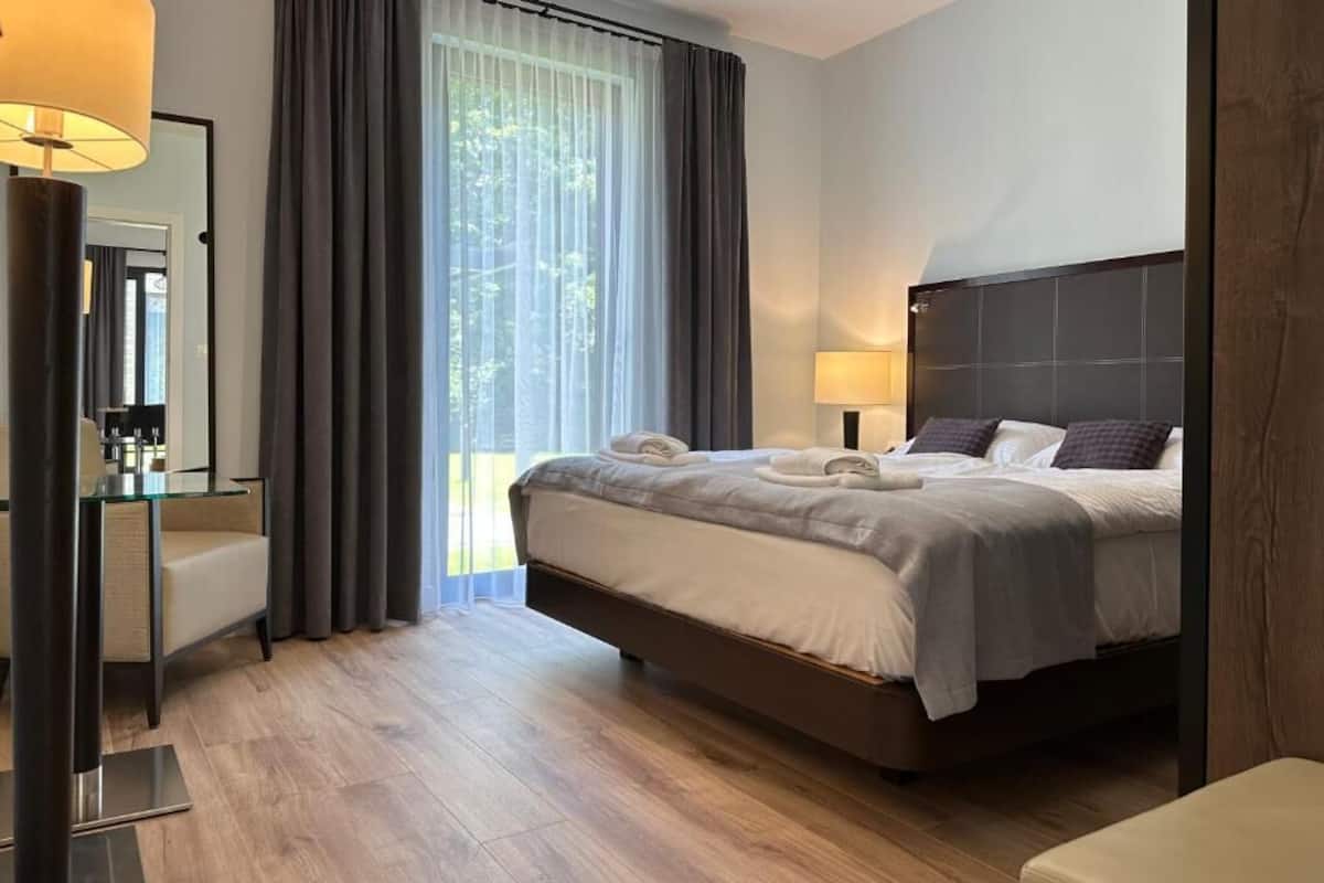 A modern hotel room with a large bed, a bedside table, a mirror, and a window with curtains.