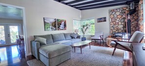 A living room with a sofa, ottoman, coffee table, and chairs. There are two windows with views of greenery, a stone wall, and a fireplace.