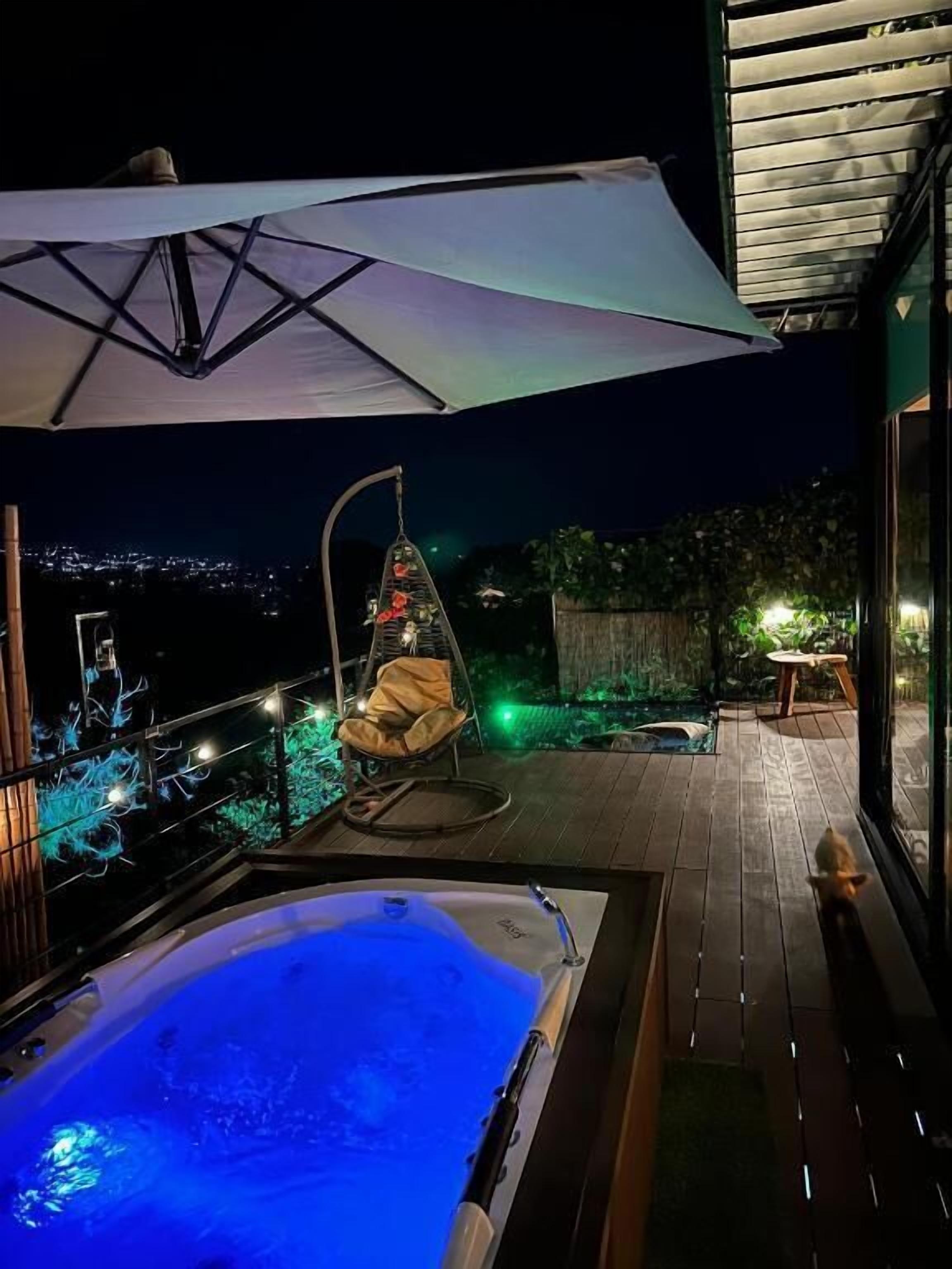 Un jacuzzi, una silla colgante y una sombrilla en una terraza de madera con vista a la ciudad de noche.