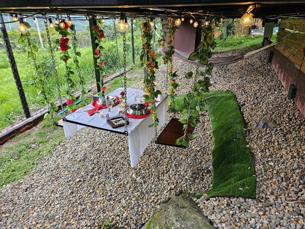 Un espace de restauration en plein air avec une table dressée pour deux personnes, entouré de galets et de verdure, sous une pergola agrémentée de guirlandes lumineuses.