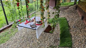 Un espace de restauration en plein air avec une table dressée pour deux personnes, entouré de galets et de verdure, sous une pergola agrémentée de guirlandes lumineuses.