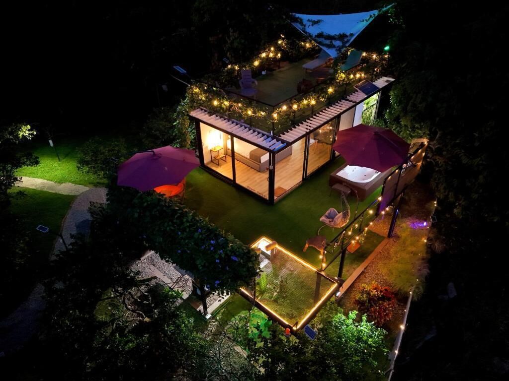 Una estructura con paredes de vidrio, un patio, mobiliario exterior e iluminación con luces colgantes.