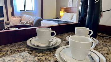 A hotel room with a bed, a bedside table with a lamp, a coffee maker, and two cups on a tray.