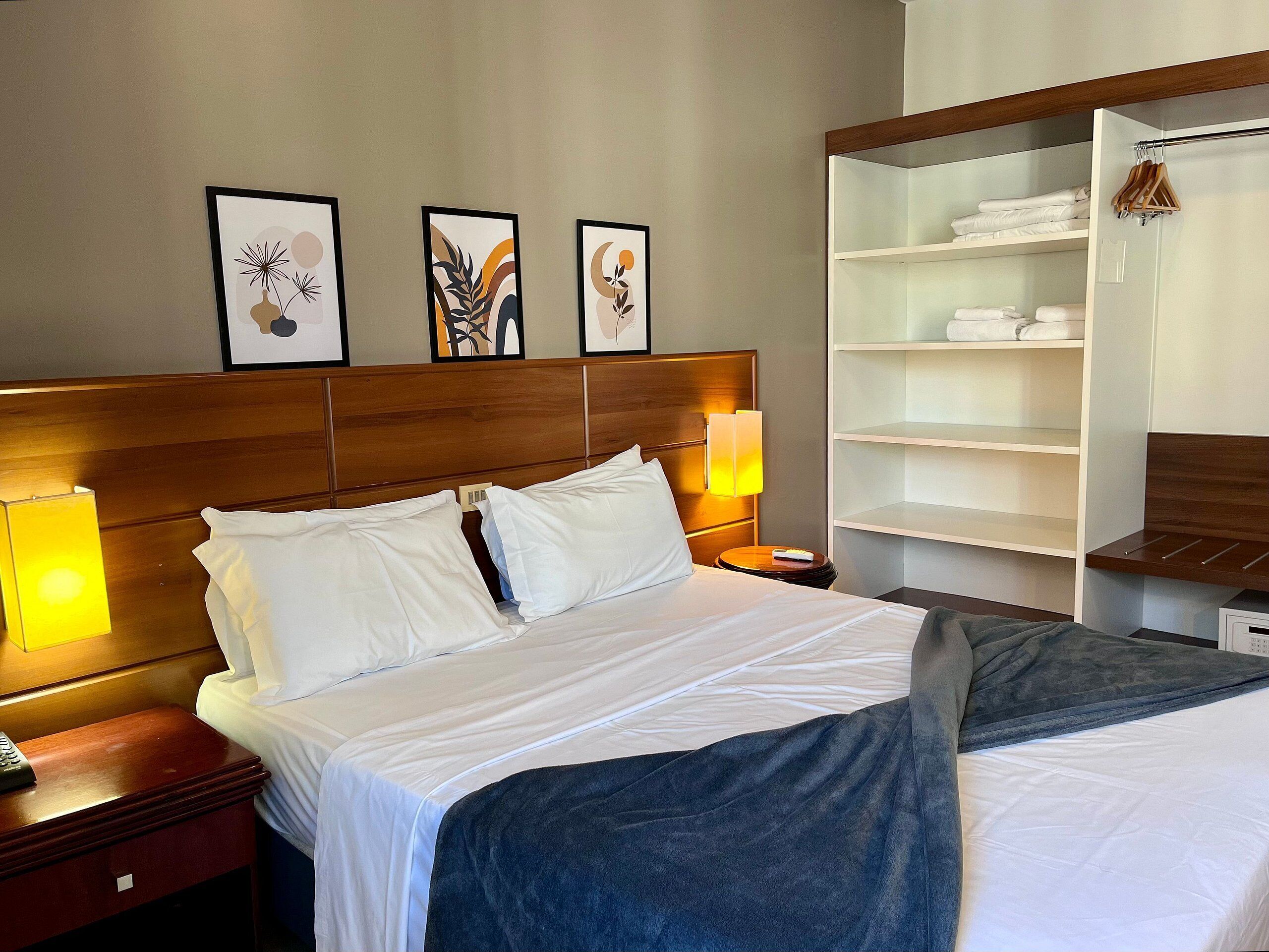 Quarto de hotel com cama, abajures, mesa de cabeceira, guarda-roupa e três quadros na parede.