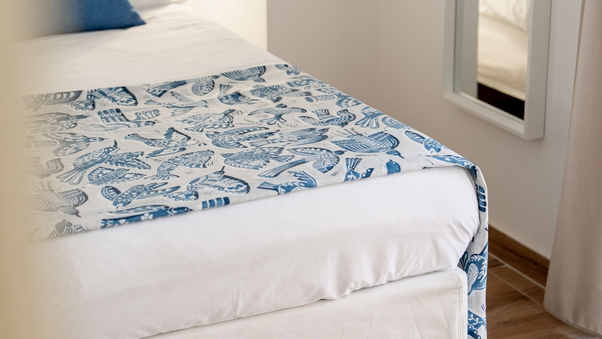 A bed with a white and blue patterned bedspread, a white pillow, and a window with a view of the outside.