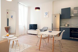Una sala de estar moderna con una televisión de pantalla plana, una mesa de comedor con sillas, una zona de cocina con refrigerador y horno, y un espejo grande.