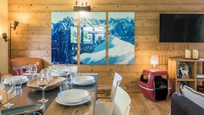 Une pièce aux murs lambrissés, avec une table à manger dressée pour quatre personnes, un panier rouge pour animaux de compagnie, une bibliothèque et un téléviseur à écran plat fixé au mur.