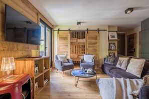 Ein gemütliches Wohnzimmer mit Flachbildfernseher, zwei Sesseln, einer Couch, einem Couchtisch und einem Bücherregal.