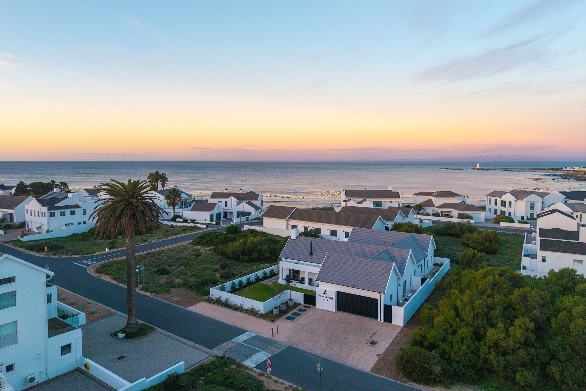 A coastal residential area with houses, a palm tree, and a view of the sea at sunset.