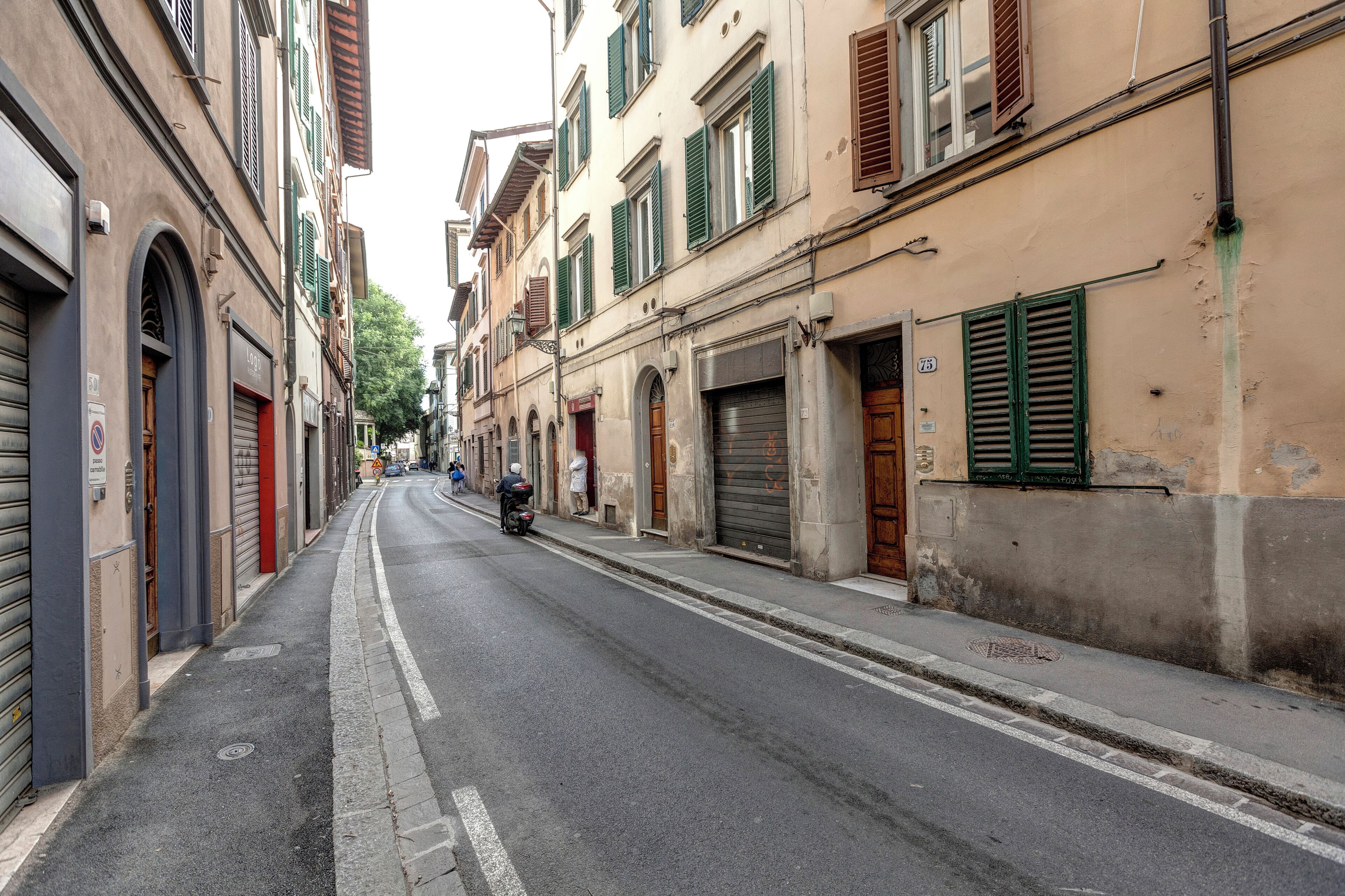 A narrow street lined with buildings, some with closed shutters, and a motorcyclist in the distance.
