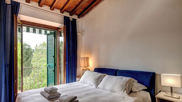 A bedroom with a bed, a nightstand, a lamp, and a view of greenery through an open door. - Romana Apartment by Mamo Florence (Florence)