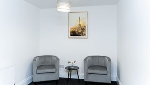 Een kamer met twee grijze fauteuils, een kleine ronde tafel met een vaas, een lijst met een foto van de Eiffeltoren en een witte plafondlamp.