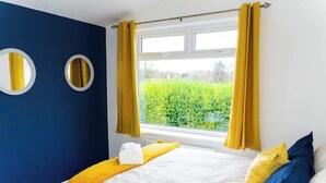 Een slaapkamer met een bed, gele gordijnen, een raam met uitzicht op groen en twee ronde spiegels aan de muur.