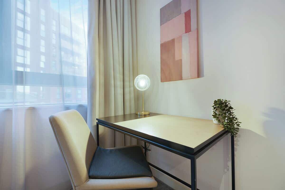 Habitación de hotel con escritorio, silla, lámpara y vistas a edificios a través del ventanal.