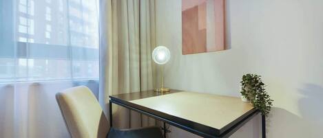 Habitación de hotel con escritorio, silla, lámpara y vistas a edificios a través del ventanal.
