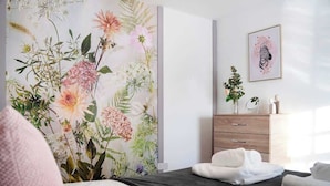 Ein Schlafzimmer mit floraler Tapete, einem Bett mit weißen Leinen, einer Holzkommode und einem gerahmten Kunstwerk an der Wand.