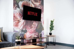 Ein Wohnzimmer mit einem an einer floralen Wand montierten Flachbildfernseher, einem grauen Sofa, einem Holztisch und einem kleinen Topfpflanzen.