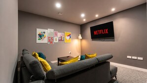 A modern living room with a gray sofa, yellow pillows, a black side table, a floor lamp, and a wall-mounted TV displaying 'NETFLIX'. - Chic Middlesbrough Studio|Sleeps 2|By Stay with us (Middlesbrough)