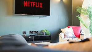 Olohuoneessa on seinälle asennettu taulutelevisio, jossa näkyy punaisilla kirjaimilla teksti »NETFLIX», punainen koristetyyny sohvan päällä, pieni kasvi sivupöydällä ja seinään kiinnitetty valaisin.