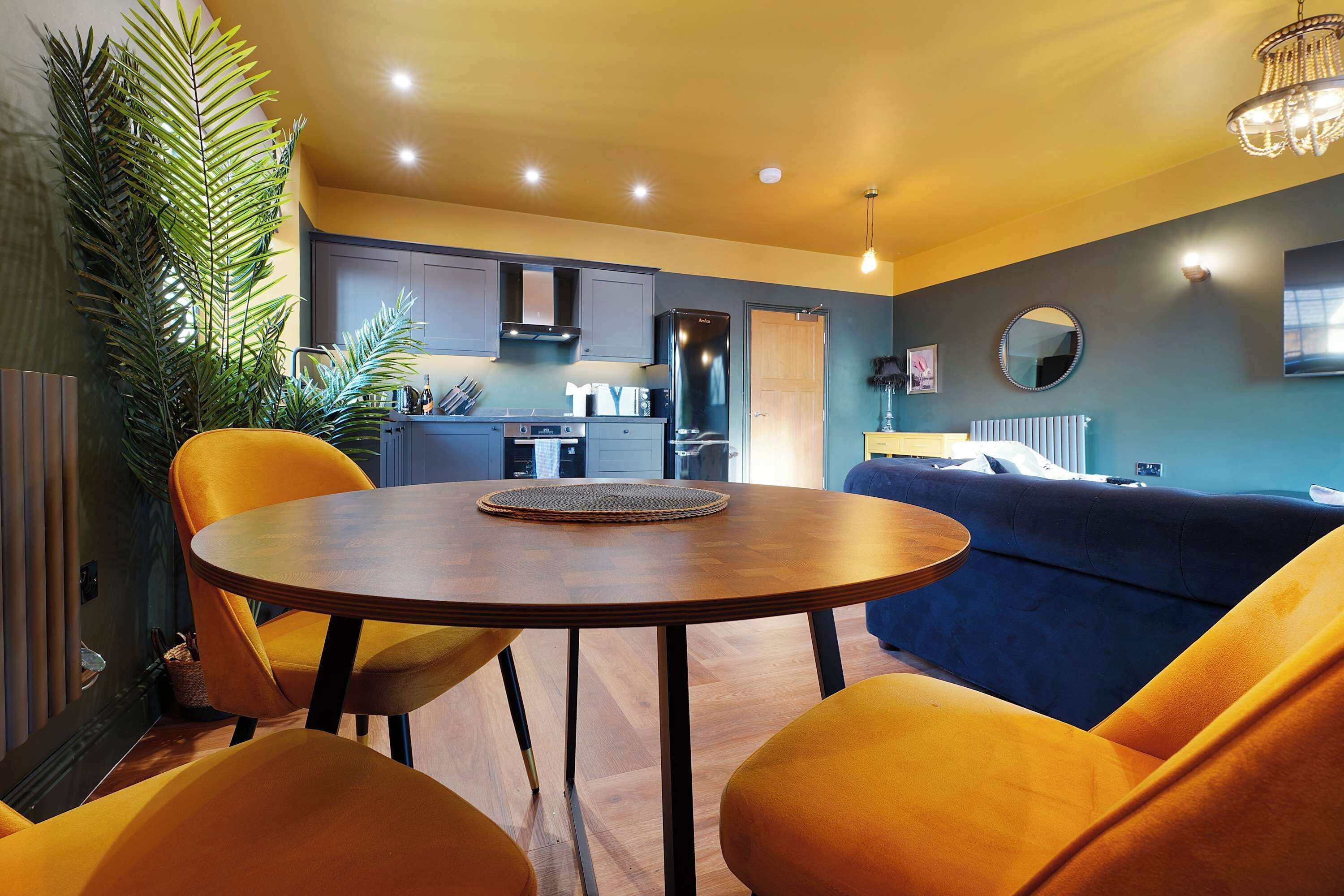 Uma sala de estar moderna com uma mesa redonda de madeira, cadeiras amarelas, um sofá azul e uma cozinha ao fundo.